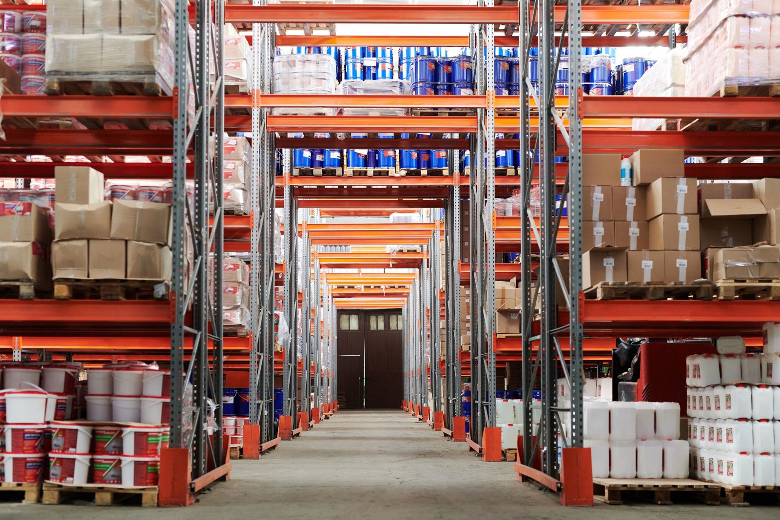 A long warehouse aisle between tall orange industrial storage racks stacked with pallets and boxes.