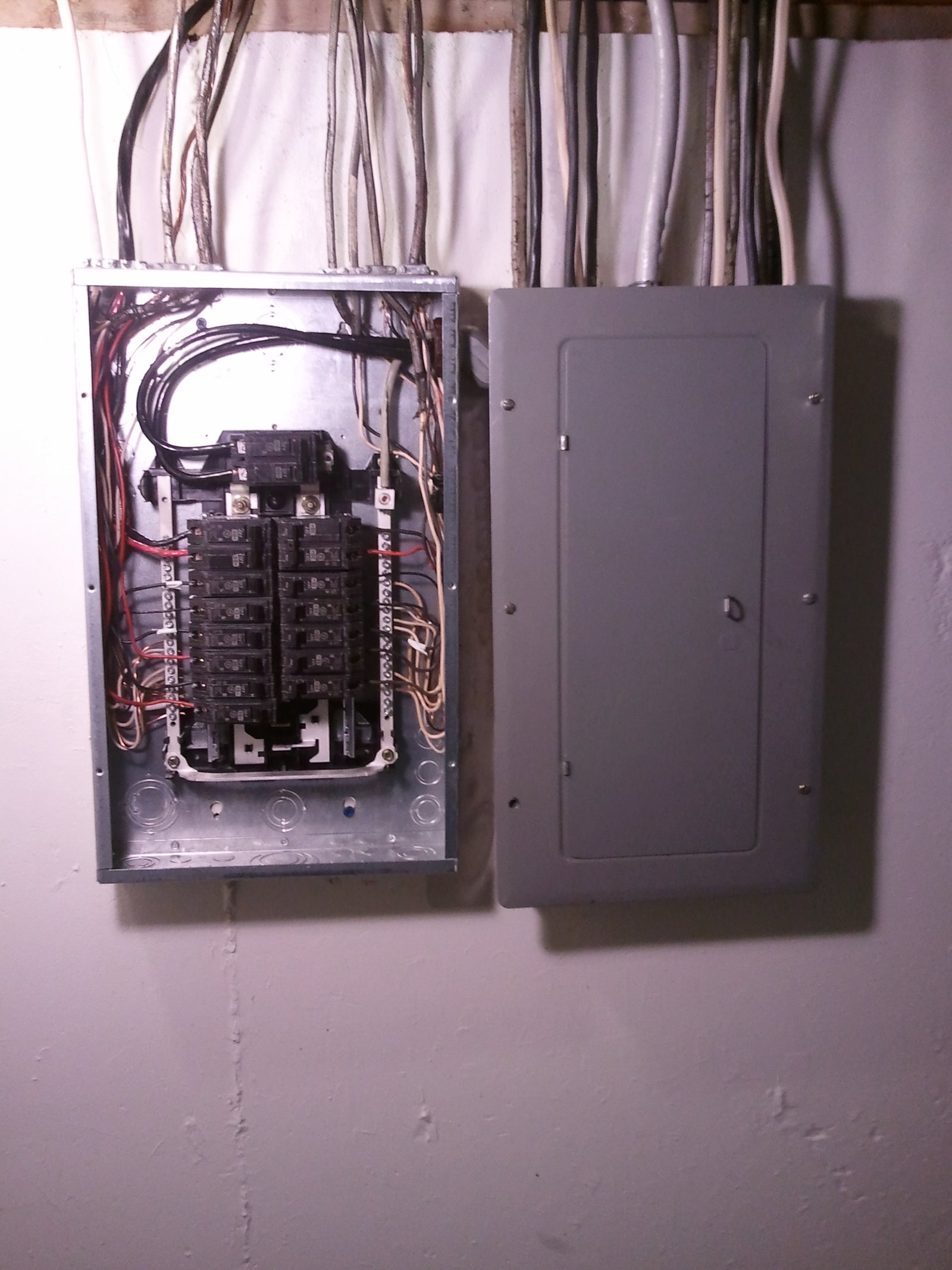 Two wall-mounted electrical panels, the upper one open with circuit breakers and wiring visible, the lower one closed.