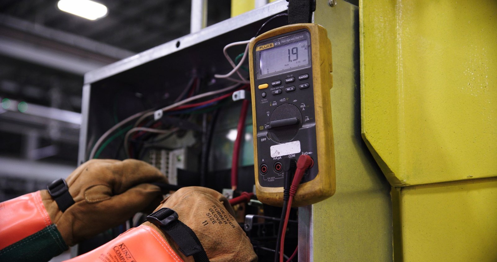 Gloved hands using a digital multimeter to test wiring inside a metal electrical enclosure.