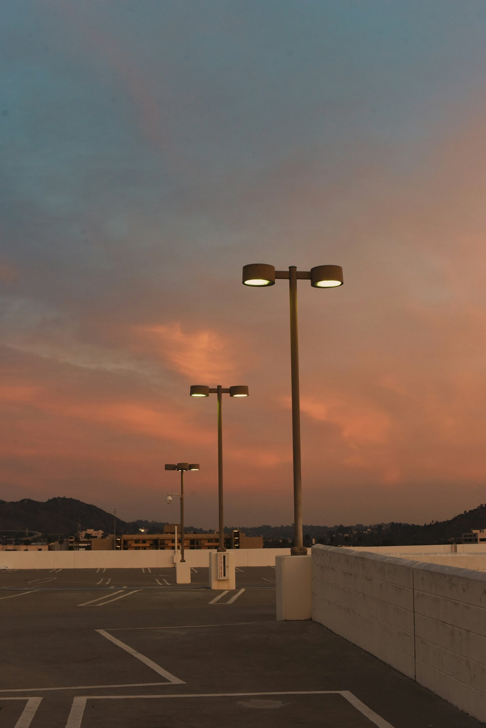 Three parking-lot light poles glowing at dusk on a rooftop deck, with hills in the distance.
